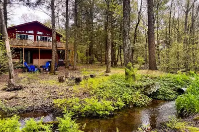 a house in the woods with a creek in front of it at Hot Tub, Dock, Pool Table! Lakefront Poconos Haven in White Haven
