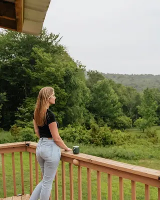 a woman standing on a wooden balcony with a cup of coffee at Rustic Escape UTV, Tub, Trails & Fire Pit 25 acre in Roscoe
