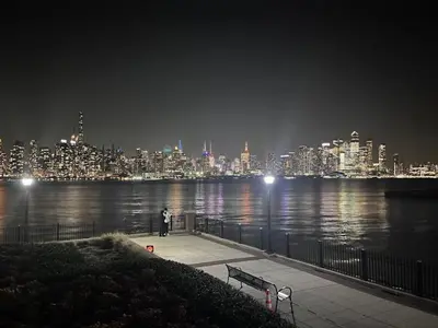 a man standing on a boardwalk near the water at night at Prime Location Explore NYC in West New York