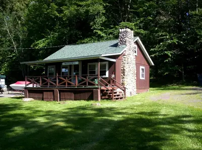 a small red house with a stone chimney in the grass at Quaint Lakefront Cabin Rental in Harveys Lake near Wilkes-Barre, Pennsylvania in Harveys Lake