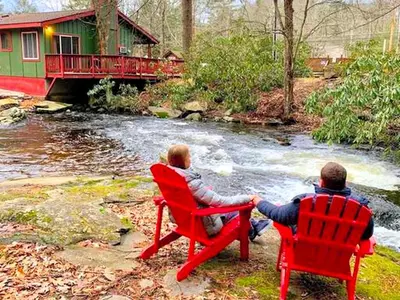 two people sitting in red chairs in front of a river at Cozy and Secluded Cabin with a Fireplace in the Poconos Mountains of Pennsylvania in Canadensis