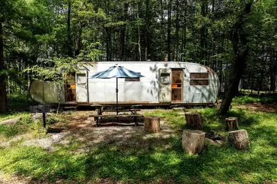 an old rv parked in the woods with an umbrella at Gorgeous Fully Restored Caravan Nestled in the Trees Along Silver Lake for a Unique Vacation in Woodridge, New York in Woodridge