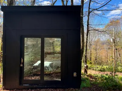 a black structure with a window in the woods at Fireplace Catskill Views Overlook Lake Cabin in Parksville