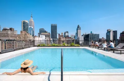 a woman in a hat sitting on the edge of a pool with a city skyline at Blueground Chelsea rooftop pool nr The Vessel NYC-2147 in New York