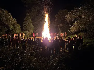 a group of people standing around a large fire at Slumberland at the River's Edge in Pond Eddy